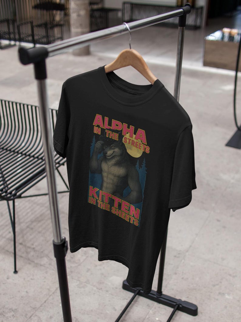 May include: Black t-shirt with a graphic of a muscular wolf-like character under a full moon. Text on the shirt reads "ALPHA IN THE STREETS, KITTEN IN THE SHEETS" in red and yellow lettering. The shirt is hanging on a wooden hanger.