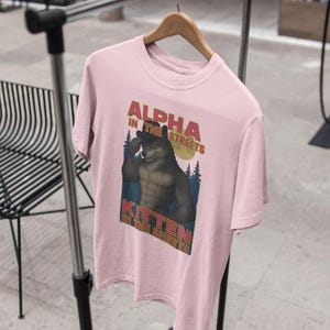 May include: A light pink t-shirt with the text "ALPHA IN THE STREETS" above an illustration of a wolf-like creature. Below the illustration is the text "KITTEN IN THE SHEETS". The shirt is hanging on a wooden hanger.