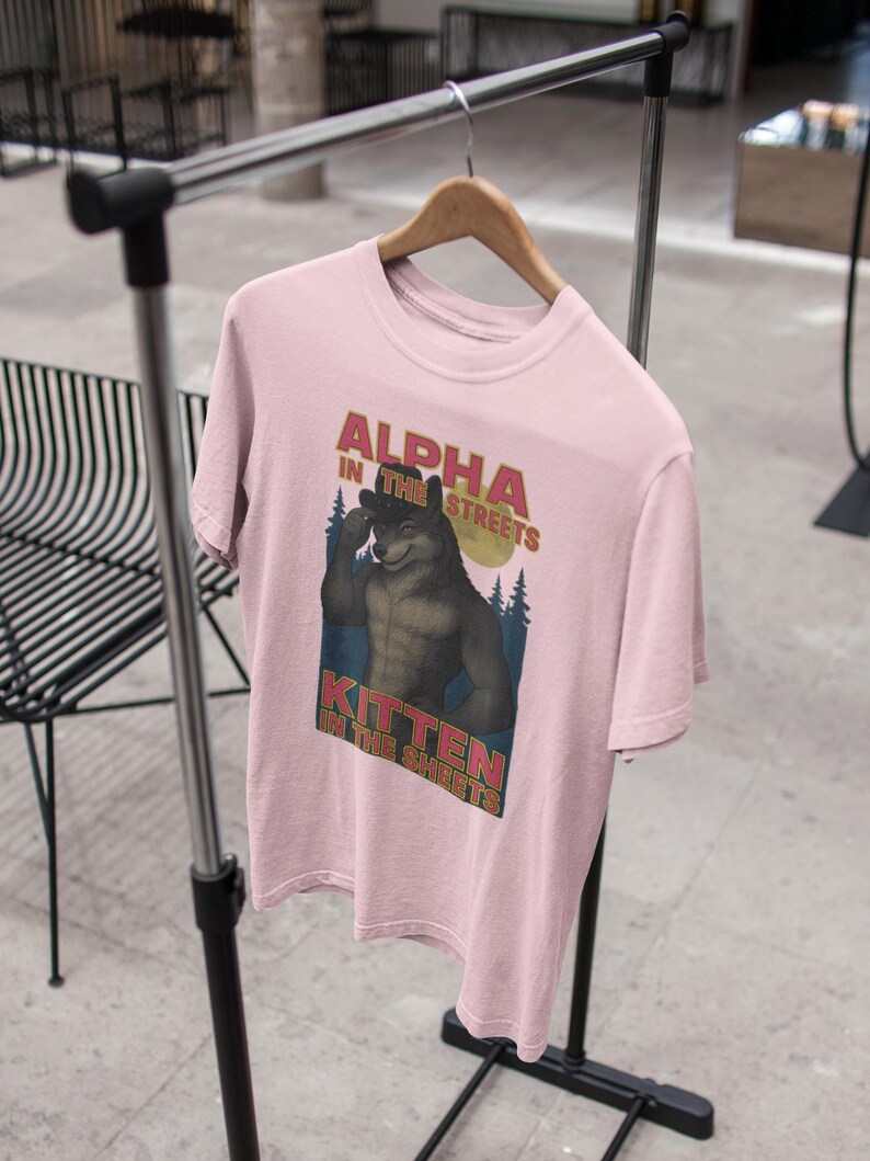 May include: A light pink t-shirt with the text "ALPHA IN THE STREETS" above an illustration of a wolf-like creature. Below the illustration is the text "KITTEN IN THE SHEETS". The shirt is hanging on a wooden hanger.
