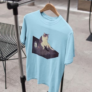 May include: Light blue t-shirt featuring a meme of a cat sitting on a dark surface with a cigarette in its mouth. The cat is white and gray. A can is next to the cat. The t-shirt is hanging on a wooden hanger.