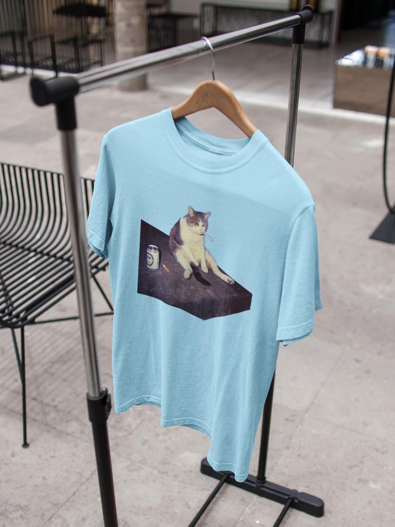 May include: Light blue t-shirt featuring a meme of a cat sitting on a dark surface with a cigarette in its mouth. The cat is white and gray. A can is next to the cat. The t-shirt is hanging on a wooden hanger.