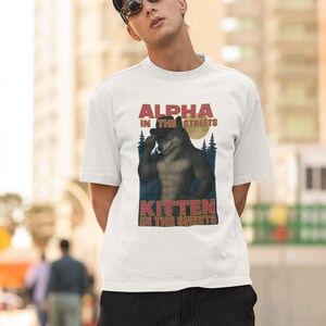 May include: White t-shirt with a graphic of a wolf-like creature with the text "ALPHA IN THE STREETS" and "KITTEN IN THE SHEETS". The shirt is worn by a person wearing a black cap and sunglasses.