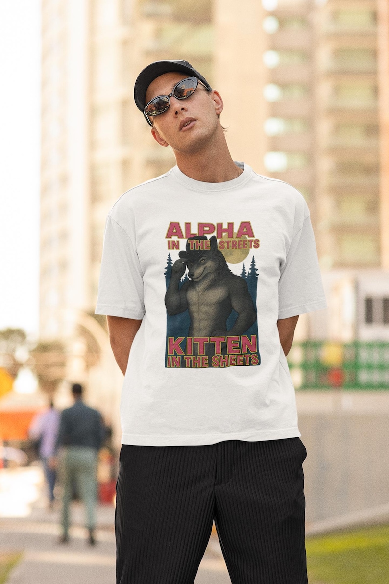 May include: White t-shirt with a graphic of a wolf-like creature with the text "ALPHA IN THE STREETS" and "KITTEN IN THE SHEETS". The shirt is worn by a person wearing a black cap and sunglasses.