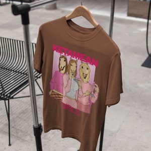 May include: Brown t-shirt with a graphic print of three women with horse heads wearing pink shirts and holding drinks. The text "Ketamine Girls" is printed on the shirt.