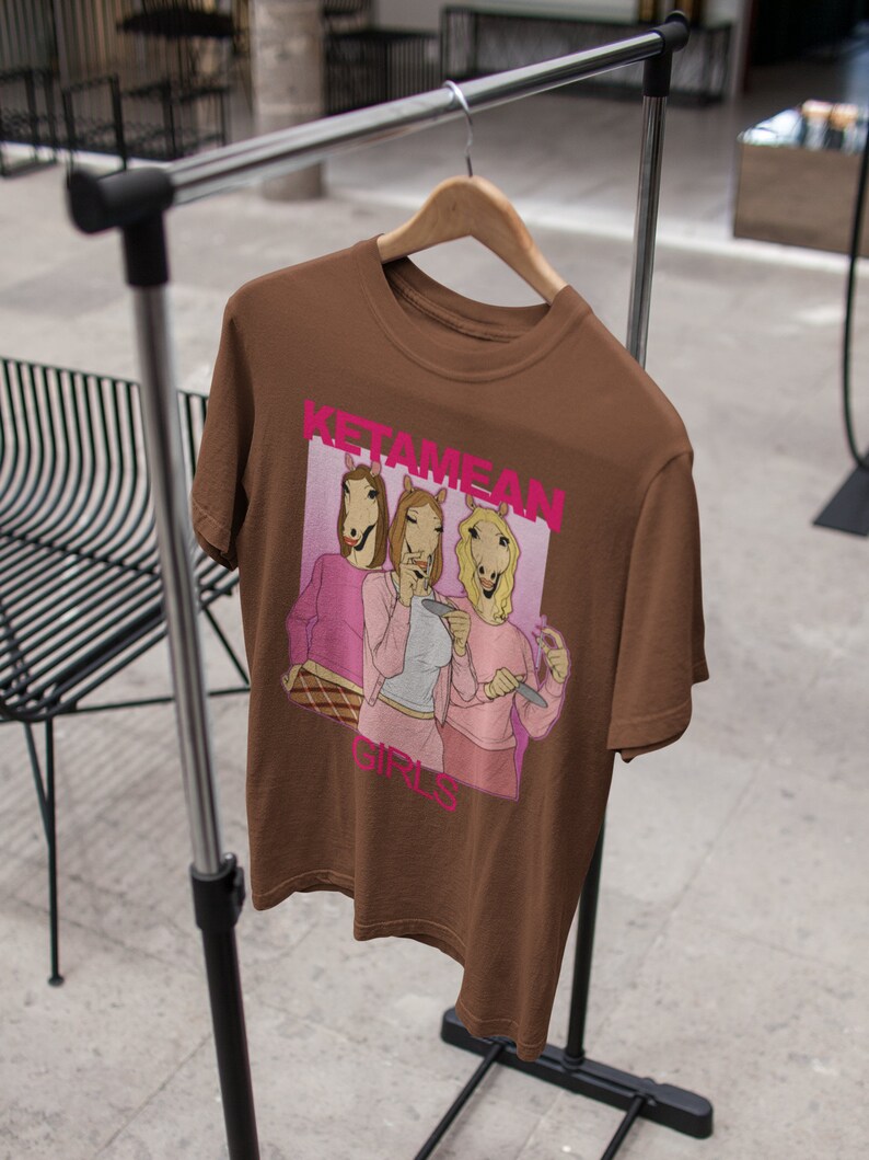 May include: Brown t-shirt with a graphic print of three women with horse heads wearing pink shirts and holding drinks. The text "Ketamine Girls" is printed on the shirt.