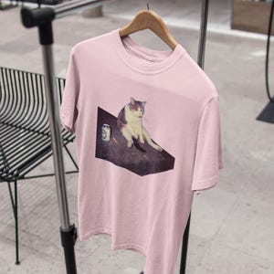 May include: A light pink t-shirt featuring a meme of a cat sitting on a table with a can. The cat is white and gray. The t-shirt is hanging on a wooden hanger on a metal clothing rack.