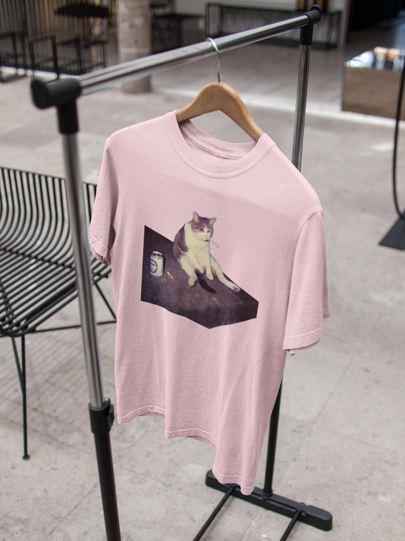 May include: A light pink t-shirt featuring a meme of a cat sitting on a table with a can. The cat is white and gray. The t-shirt is hanging on a wooden hanger on a metal clothing rack.