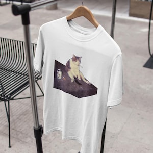 May include: White t-shirt featuring a meme of a cat sitting with a cigarette and a can. The cat is depicted in shades of gray and white, sitting on a dark surface. The t-shirt is hanging on a clothing rack.