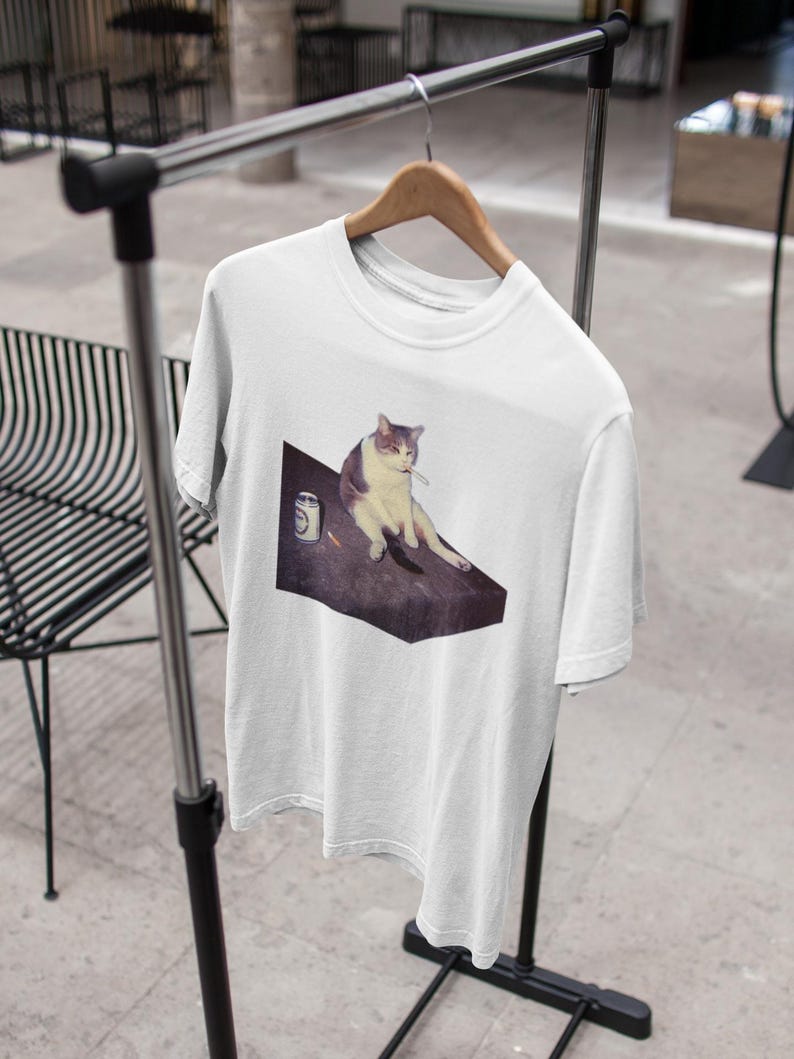 May include: White t-shirt featuring a meme of a cat sitting with a cigarette and a can. The cat is depicted in shades of gray and white, sitting on a dark surface. The t-shirt is hanging on a clothing rack.