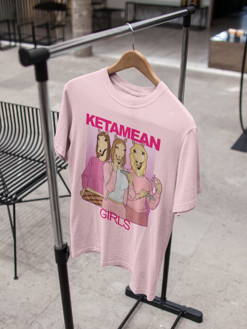 May include: Pink t-shirt with a graphic of three women with horse heads wearing pink outfits and the text "Ketamine Girls".