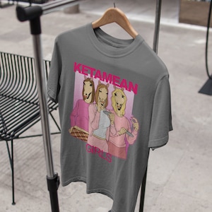 May include: Gray t-shirt with a graphic of three women with horse heads wearing pink shirts and holding drinks. The text "Ketamine Girls" is printed in pink on the shirt.