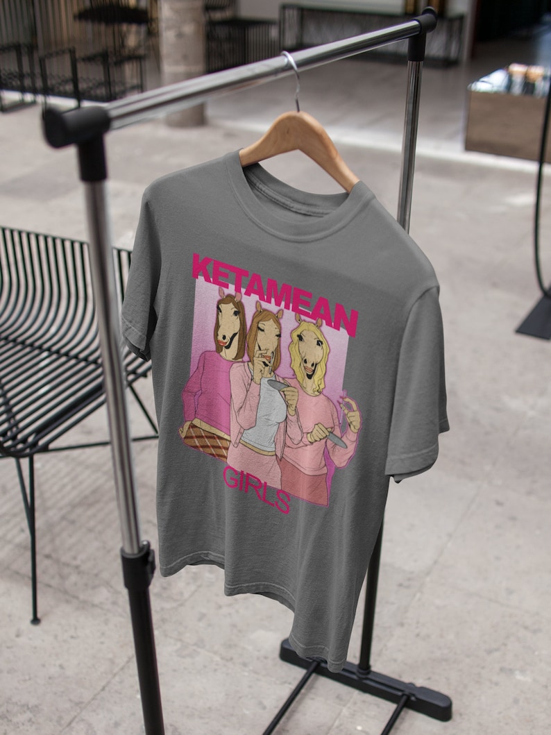 May include: Gray t-shirt with a graphic of three women with horse heads wearing pink shirts and holding drinks. The text "Ketamine Girls" is printed in pink on the shirt.