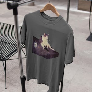 May include: Dark gray t-shirt featuring a graphic of a cat with a cigarette in its mouth, sitting on a table next to a can. The cat is white and gray. The t-shirt is hanging on a wooden hanger.
