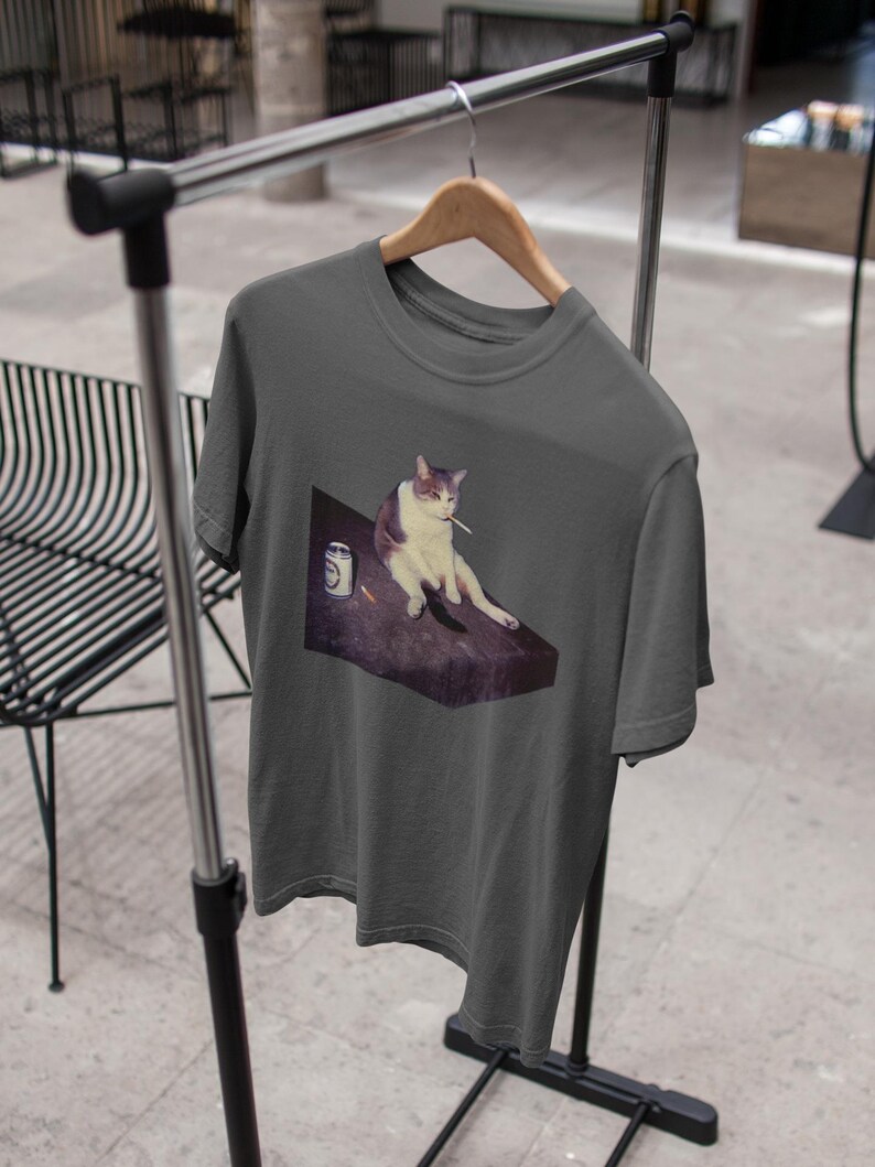 May include: Dark gray t-shirt featuring a graphic of a cat with a cigarette in its mouth, sitting on a table next to a can. The cat is white and gray. The t-shirt is hanging on a wooden hanger.