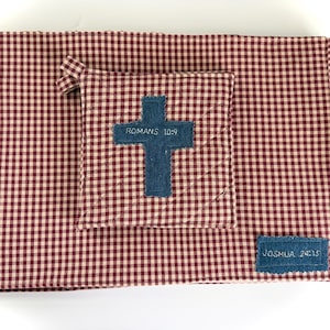 May include: A rectangular fabric mat and a smaller square quilted potholder, both featuring a red and white gingham pattern. The potholder displays a blue fabric cross with the text ROMANS 10:9, and the mat has a blue fabric label with JOSHUA 24:15.