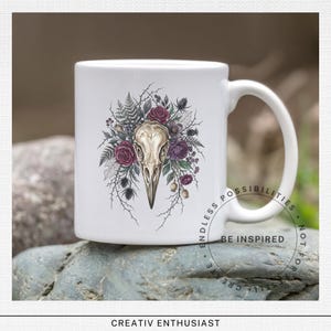 White mug with crow skull and floral design, "Be Inspired" text.