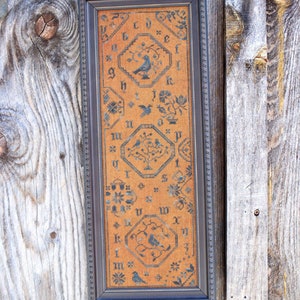 May include: Framed cross-stitch sampler with the text "Quaker Blue Bird" at the top. The sampler features blue bird and floral designs on an orange background, framed in a dark gray frame. The website address "www.willowhillsamplings.net" is at the bottom.