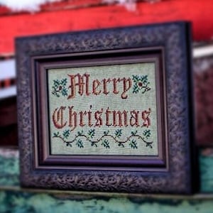 Può includere: Un ricamo a punto croce incorniciato con uno sfondo bianco e ricamo verde. Il ricamo recita "Merry Christmas" in un carattere rosso e corsivo.