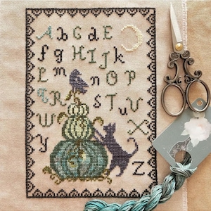 May include: Cross-stitch embroidery of an alphabet with a black cat and a stack of pumpkins. The letters are in a traditional font and the pumpkins are green and white. The cat is black and has a white face. The background is a light beige fabric.