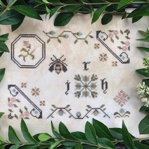 May include: A white cross-stitch sampler featuring a bee, flowers, and the letters 'J' and 'H' in a brown, green, and blue colour scheme.