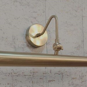 May include: A brass-toned picture light fixture. The fixture has a circular wall mount, a curved arm, and a cylindrical light bar. The finish is a warm, metallic gold color, suitable for illuminating artwork or decor.