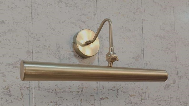 May include: A brass-toned picture light fixture. The fixture has a circular wall mount, a curved arm, and a cylindrical light bar. The finish is a warm, metallic gold color, suitable for illuminating artwork or decor.