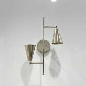 Puede incluir: Una lámpara de pared plateada con dos apliques ajustables. La lámpara tiene una base redonda y un solo brazo que se extiende hacia un lado. Los apliques tienen forma de cono y tienen un acabado plateado.