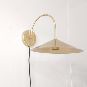 May include: A brass wall sconce with a curved arm and a conical shade. The fixture is mounted on a round base and has a black power cord. The sconce is a warm gold color and is mounted on a white wall.