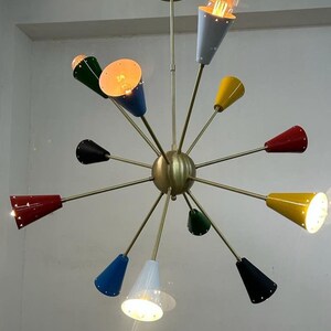 Może przedstawiać: Żyrandol w stylu mid-century modern z mosiężnym wykończeniem i wieloma stożkowymi kloszami w r&oacute;żnych kolorach, w tym czerwonym, ż&oacute;łtym, niebieskim, zielonym i czarnym. Oprawy oświetleniowe są podświetlone.