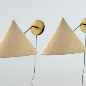 May include: A pair of wall-mounted lamps featuring beige conical fabric shades. Each lamp has a circular brass wall plate and a thin, angled brass arm connecting the shade to the base. A thin black power cord hangs down from each brass wall plate.