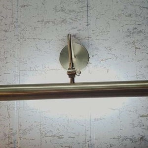 May include: A brushed nickel picture light fixture. The light has a cylindrical bar with a bright light shining from it. The fixture is mounted on a round base with an adjustable arm.