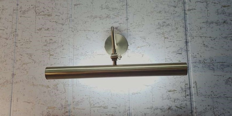 May include: A brushed nickel picture light fixture. The light has a cylindrical bar with a bright light shining from it. The fixture is mounted on a round base with an adjustable arm.