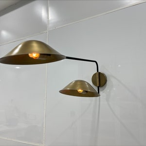 May include: A gold wall sconce with two shades. The shades are round and have a brass finish. The sconce is mounted on a white wall.