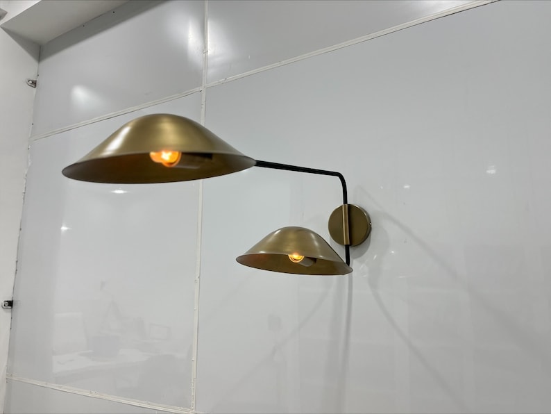May include: A gold wall sconce with two shades. The shades are round and have a brass finish. The sconce is mounted on a white wall.