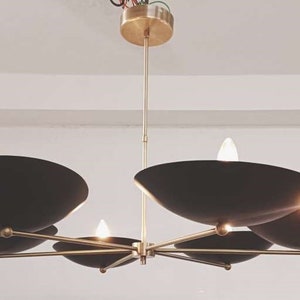 May include: A contemporary chandelier featuring a brass frame and six black, bowl-shaped shades. The light fittings are arranged around a central point, with small light bulbs providing illumination. The chandelier is suspended from the ceiling.
