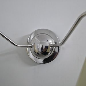 Puede incluir: Aplique de pared cromado pulido con dos brazos curvos que se extienden desde una base circular. La base refleja el entorno. El aplique est&aacute; montado en una pared blanca y se ve una pantalla de l&aacute;mpara color crema.