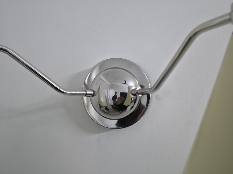 Puede incluir: Aplique de pared cromado pulido con dos brazos curvos que se extienden desde una base circular. La base refleja el entorno. El aplique est&aacute; montado en una pared blanca y se ve una pantalla de l&aacute;mpara color crema.