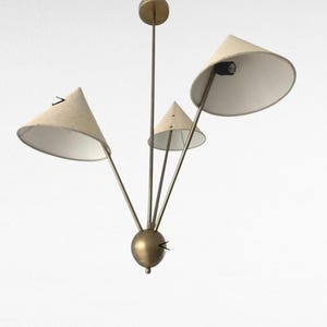 May include: A mid-century modern chandelier with three cone-shaped shades. The shades are a light beige colour, and the metal frame has a brass finish. The fixture is designed to be mounted on a ceiling.