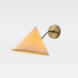 Könnte beinhalten: Eine Wandleuchte mit einem dreieckigen, kegelförmigen Schirm aus warmem, strukturiertem beigem Stoff. Der Schirm ist mit einem dünnen, abgewinkelten Metallarm an einer runden Wandplatte aus gebürstetem Messing befestigt, was eine gerichtete Leuchte ergibt.