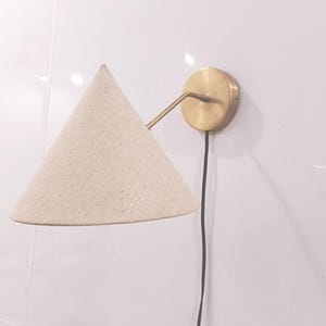May include: A wall-mounted light fixture with a conical, off-white fabric shade. The fixture has a brass-colored arm and circular base, and a black power cord. The light is mounted on a white wall.