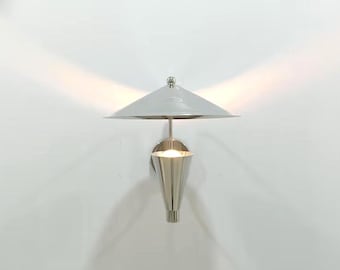 May include: A wall-mounted light fitting featuring a polished silver-tone conical base and a wide, flat, circular shade in a matching metallic finish. The lamp emits a bright, focused glow from the centre, casting light upwards and downwards.