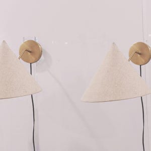 Könnte beinhalten: Zwei Wandleuchten mit konischen, cremefarbenen Stoffschirmen. Jede Leuchte hat eine kreisförmige, messingfarbene Wandhalterung und ein schwarzes Netzkabel. Die Lampen sind an einer weißen Wand montiert.