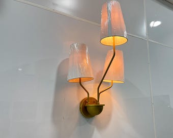 May include: A gold wall sconce with three white fabric lampshades. The sconce has a curved design with the lampshades extending out from the wall.