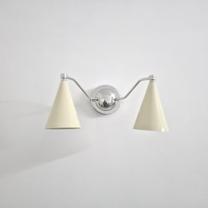 Puede incluir: Una moderna l&aacute;mpara de pared con dos pantallas c&oacute;nicas de color crema. La l&aacute;mpara tiene un acabado cromado pulido, incluyendo la placa de montaje central y los brazos curvos. La luz est&aacute; dise&ntilde;ada para uso en interiores.