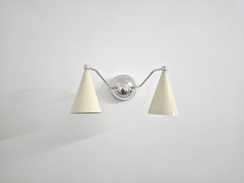 Puede incluir: Una moderna l&aacute;mpara de pared con dos pantallas c&oacute;nicas de color crema. La l&aacute;mpara tiene un acabado cromado pulido, incluyendo la placa de montaje central y los brazos curvos. La luz est&aacute; dise&ntilde;ada para uso en interiores.