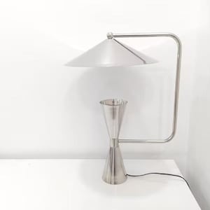 Könnte beinhalten: Eine moderne Schreibtischlampe in Silberoptik mit konischem Schirm und einer Sanduhr-förmigen Basis. Die Lampe hat einen gebogenen Arm und ein Netzkabel. Die Lampe steht auf einer weißen Oberfläche.