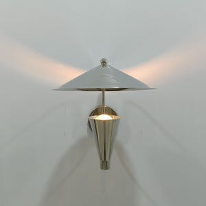 Puede incluir: Aplique de pared moderno con una pantalla gris en forma de paraguas y una lámpara cónica plateada pulida. El aplique está montado en una pared blanca y la luz proyecta un brillo cálido.