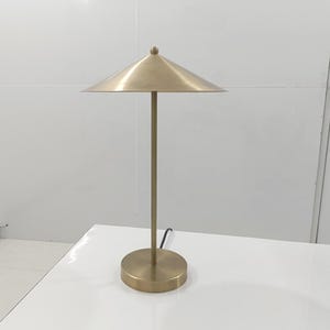 Könnte beinhalten: Messingfarbene Tischlampe mit konischem Schirm und rundem Fuß. Die Lampe hat einen schlanken Stiel und einen kleinen, dekorativen Knauf. Die Lampe steht auf einer weißen Oberfläche. Höhe ca. 50 cm.