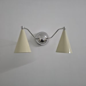Puede incluir: Una l&aacute;mpara de pared con dos pantallas c&oacute;nicas de color crema. La l&aacute;mpara tiene un acabado cromado pulido, incluyendo la placa de montaje central y los brazos curvos que sostienen las pantallas. La luz est&aacute; montada en una pared blanca.