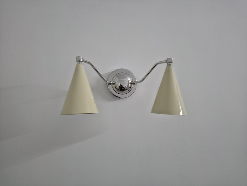 Puede incluir: Una l&aacute;mpara de pared con dos pantallas c&oacute;nicas de color crema. La l&aacute;mpara tiene un acabado cromado pulido, incluyendo la placa de montaje central y los brazos curvos que sostienen las pantallas. La luz est&aacute; montada en una pared blanca.